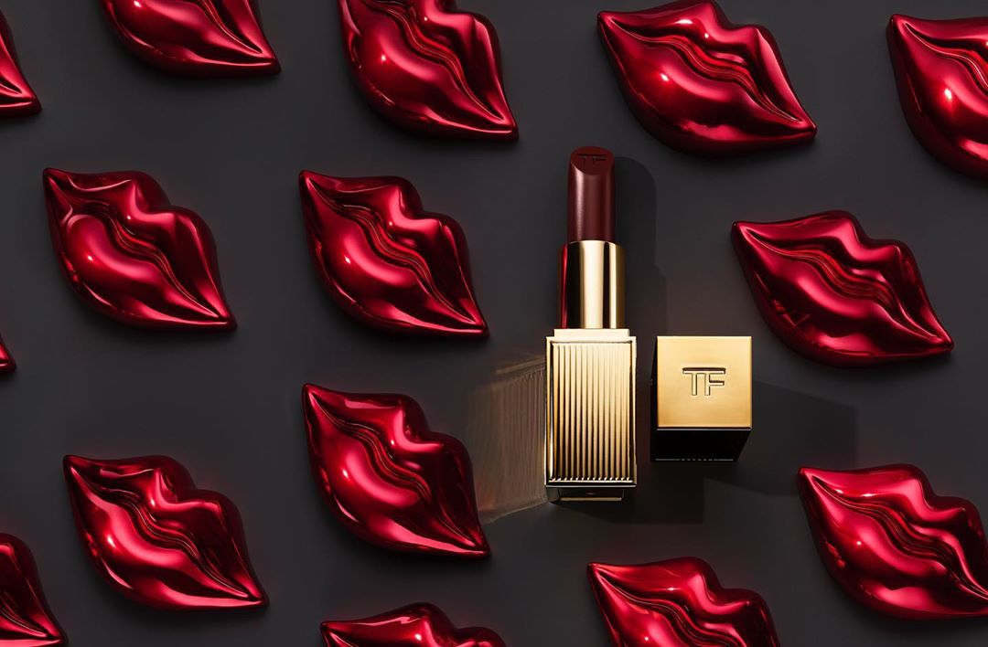 Tom Ford lipstick tube and square container with logo next to shiny red lip-shaped objects on a grey surface in a studio shot.