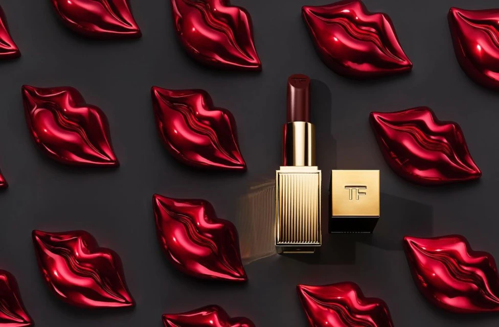 Tom Ford lipstick tube and square container with logo next to shiny red lip-shaped objects on a grey surface in a studio shot.