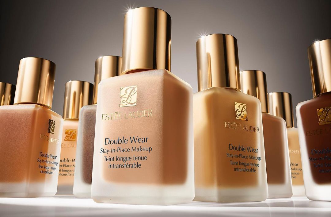 Several Estee Lauder Double Wear Stay-in-Place Makeup foundation bottles with gold lids in neutral and brown shades are presented.