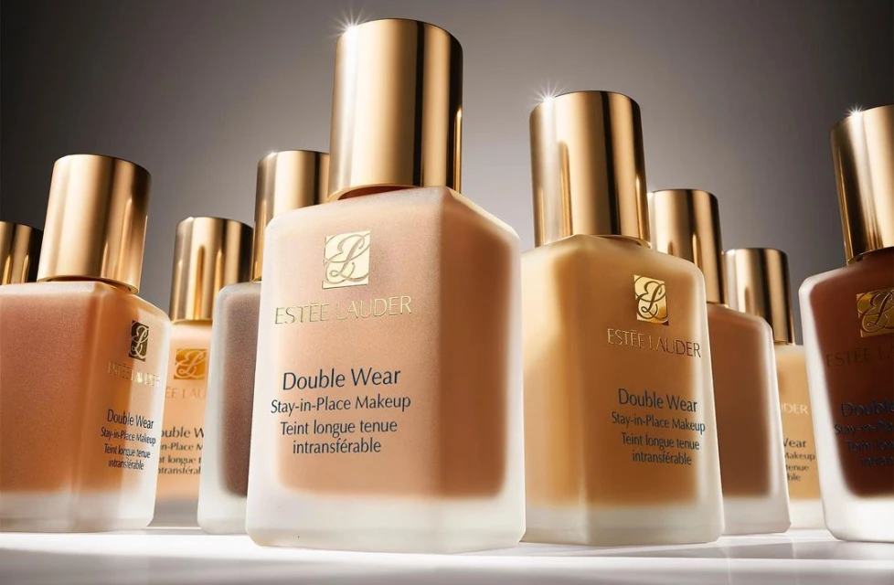 Several Estee Lauder Double Wear Stay-in-Place Makeup foundation bottles with gold lids in neutral and brown shades are presented.