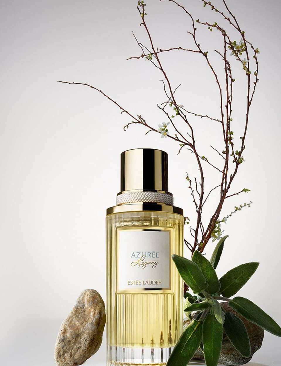 Close-up studio shot of Azurée Legacy perfume bottle, gold cap, branches, sage leaf, and stone against a clean background.