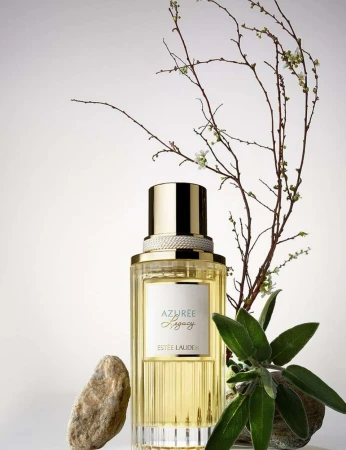Close-up studio shot of Azurée Legacy perfume bottle, gold cap, branches, sage leaf, and stone against a clean background.
