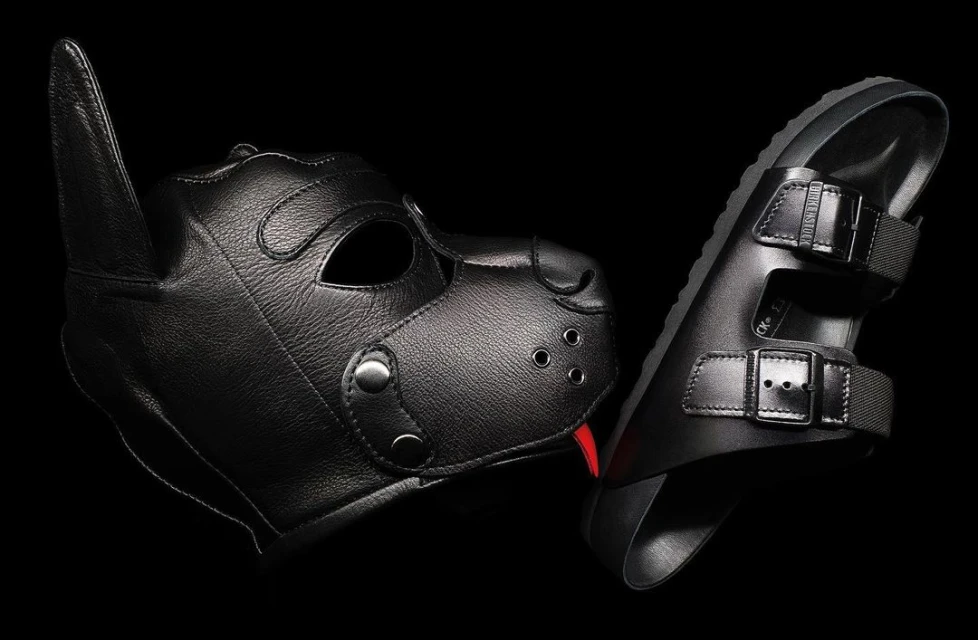 Black leather dog mask with a red tongue sticking out, next to black Birkenstock-style sandals on a black background.
