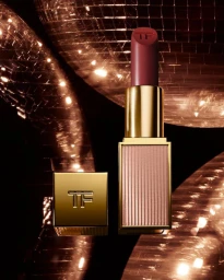 Tom Ford lipstick in a gold case with its cap removed against a reflective dark background with warm bokeh lights.