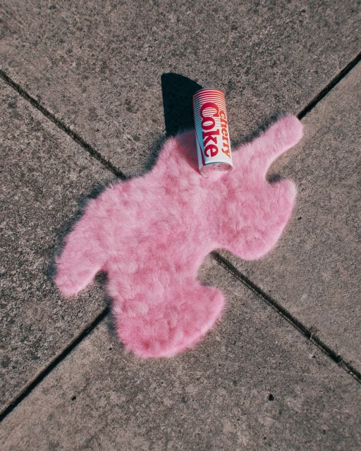 Overhead shot of a can of Cherry Coke on a pink faux fur rug placed on gray concrete with dark lines, suggesting an outdoor setting.