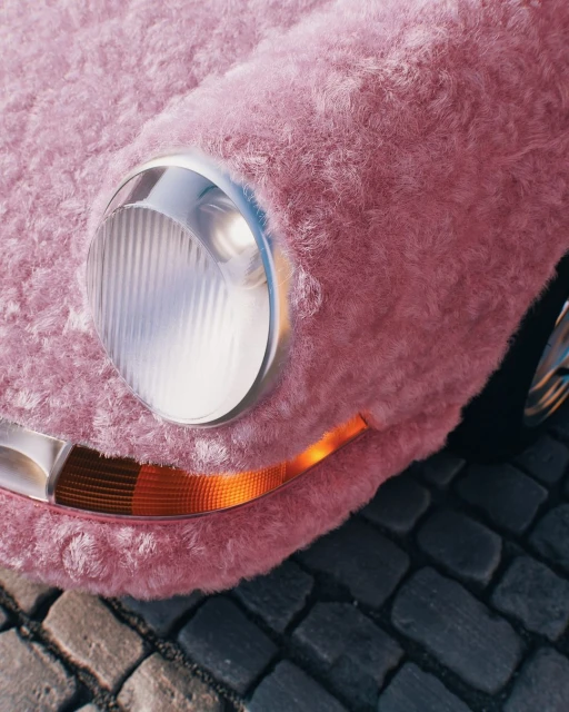 Close-up of a vintage car covered in pink fur, headlight and turn signal visible, parked on a cobblestone street in daylight.