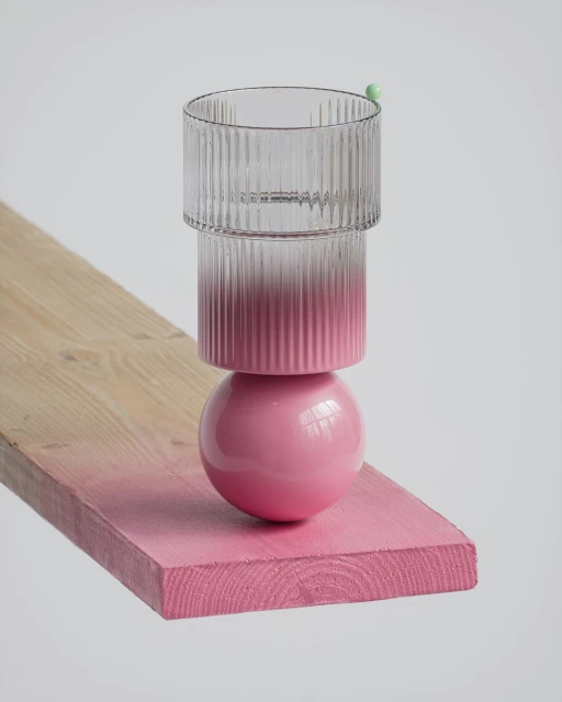 Clear ribbed glass sits atop a pink sphere on a pink-stained wooden plank against a white background, minimalist still life.