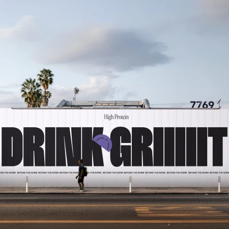 A pedestrian walks past a large street billboard displaying bold black typography reading Drink Griiiit High Protein against an open sky.