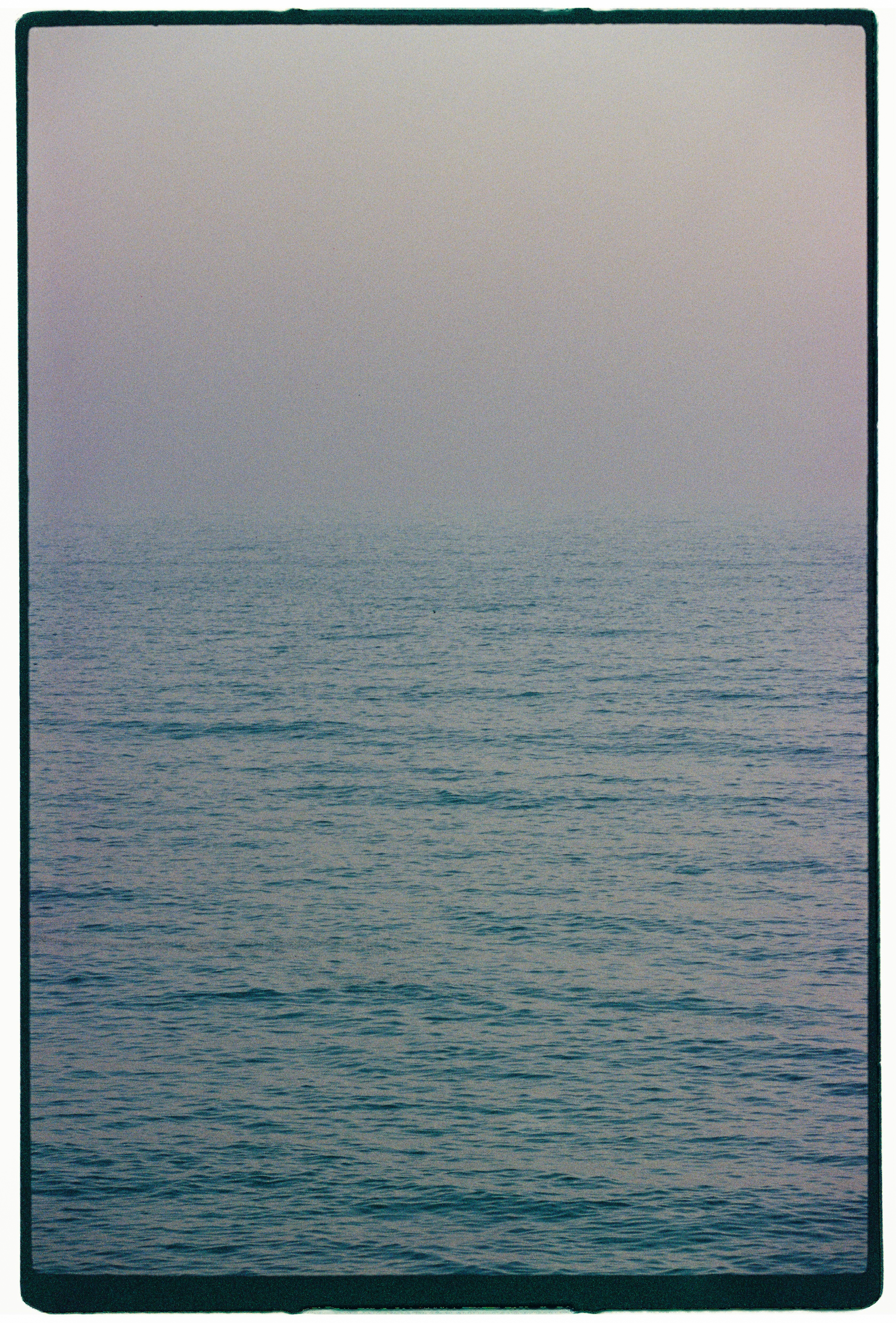 Minimalist seascape photograph showing calm blue ocean waters fading into a misty, pale overcast horizon with a dark film border.