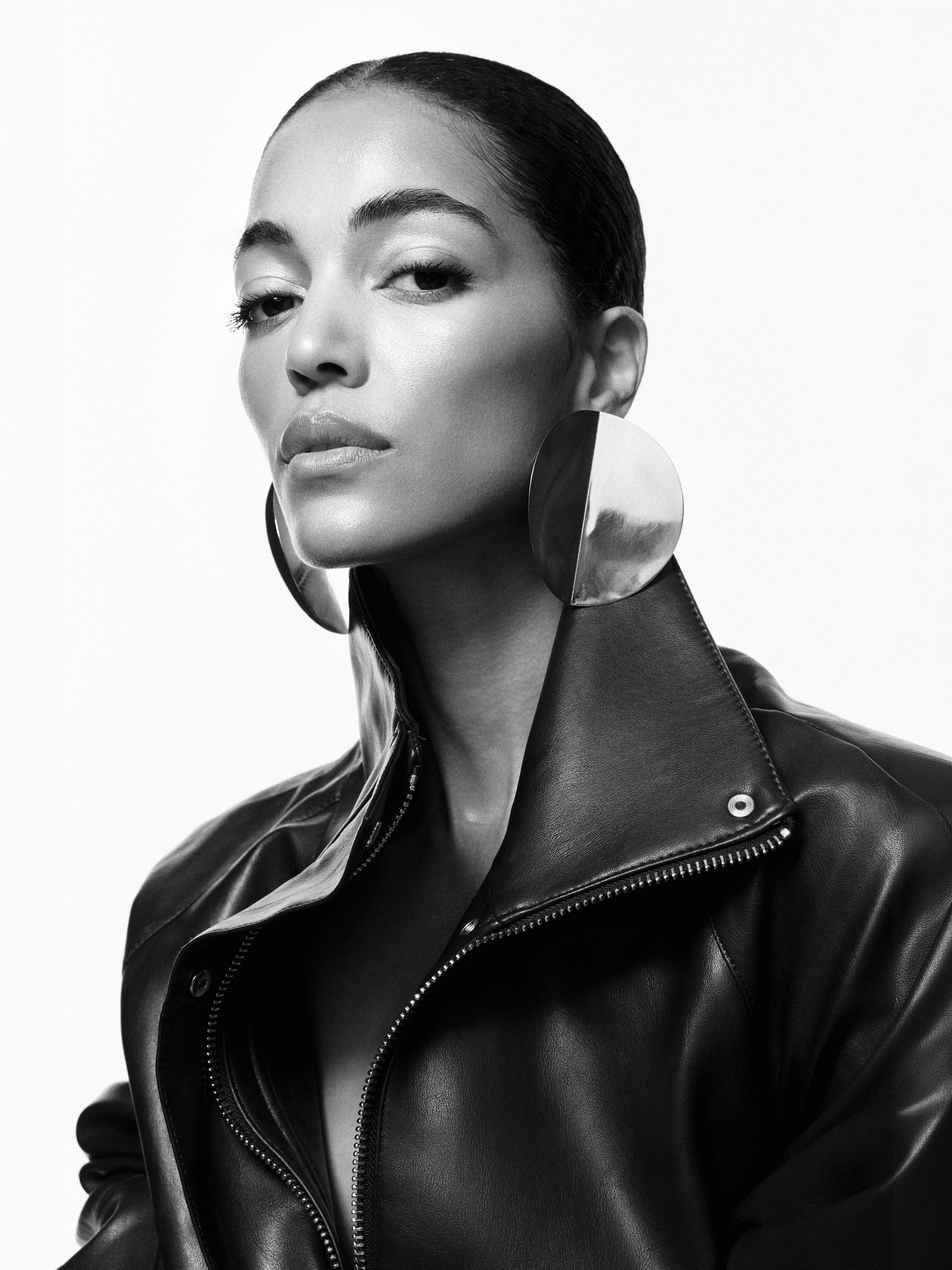 Close-up portrait of a woman with slicked-back hair wearing large circular metallic earrings and a black leather jacket against white background.