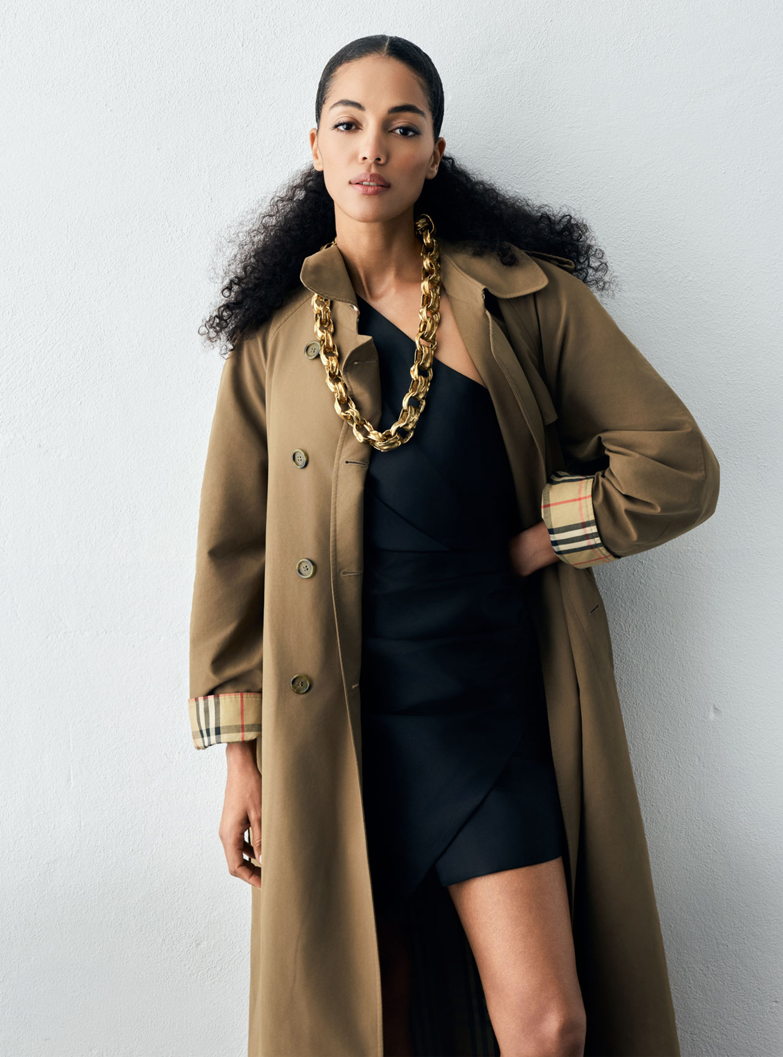A woman with dark curly hair wearing an olive trench coat over a black one-shoulder dress, accessorized with a thick gold chain necklace.