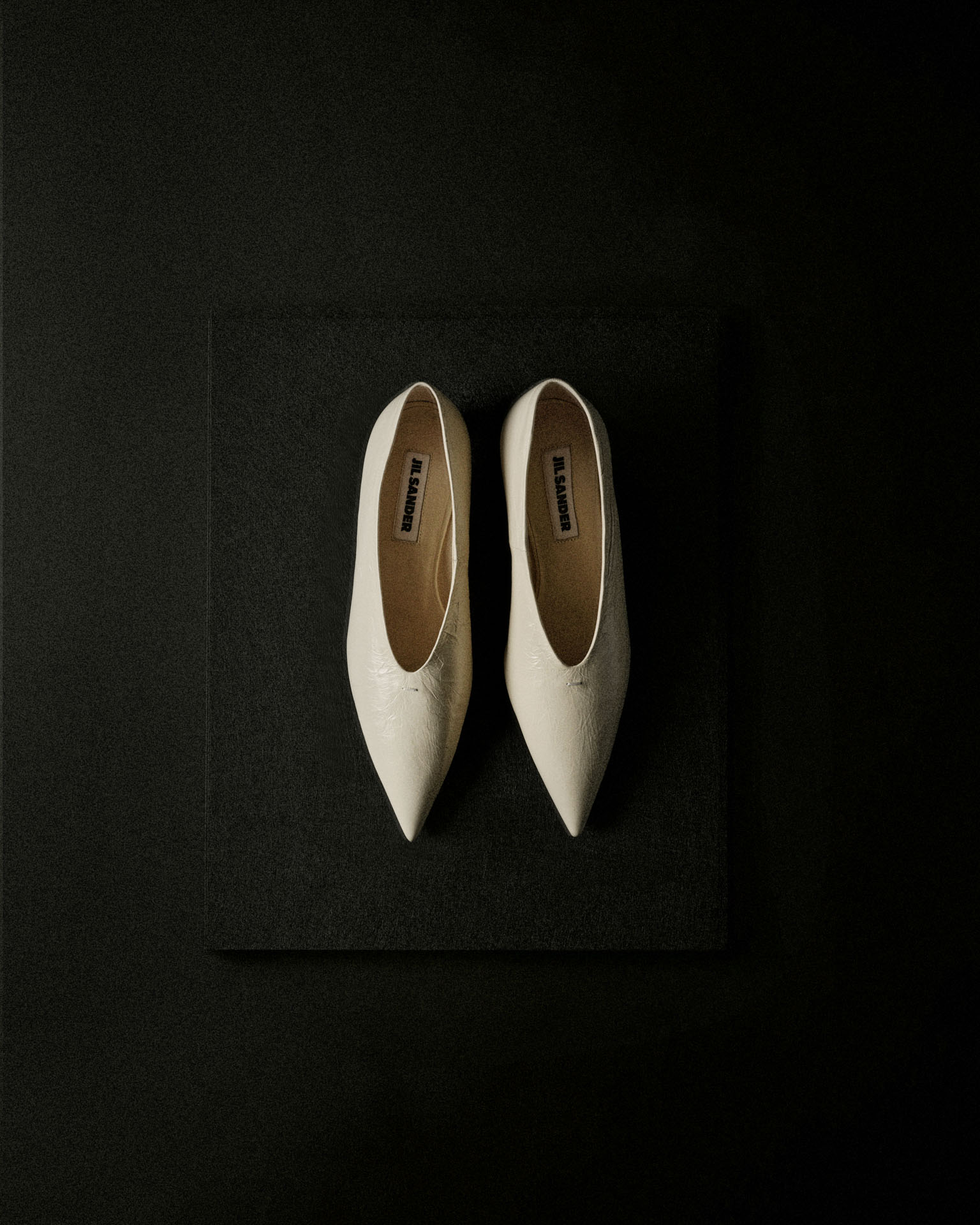 A pair of white, pointed-toe Jil Sander flats resting on a rectangular, black, textured stone base against a dark background.
