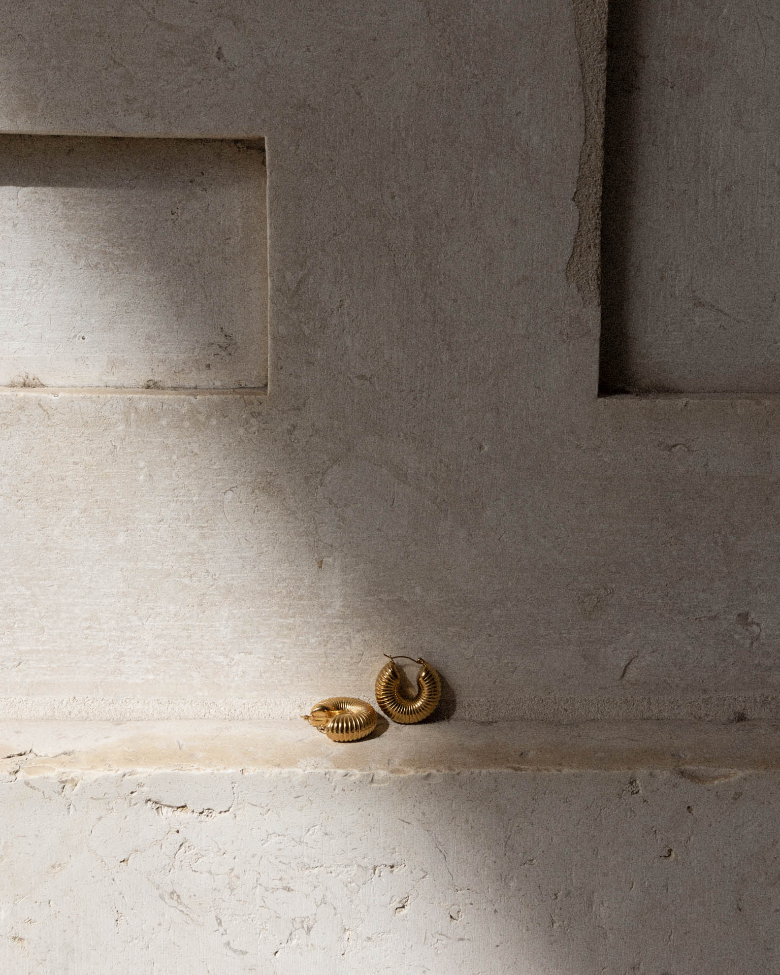 Pair of chunky ribbed gold hoop earrings resting on a rough stone wall ledge with dramatic diagonal shadow.