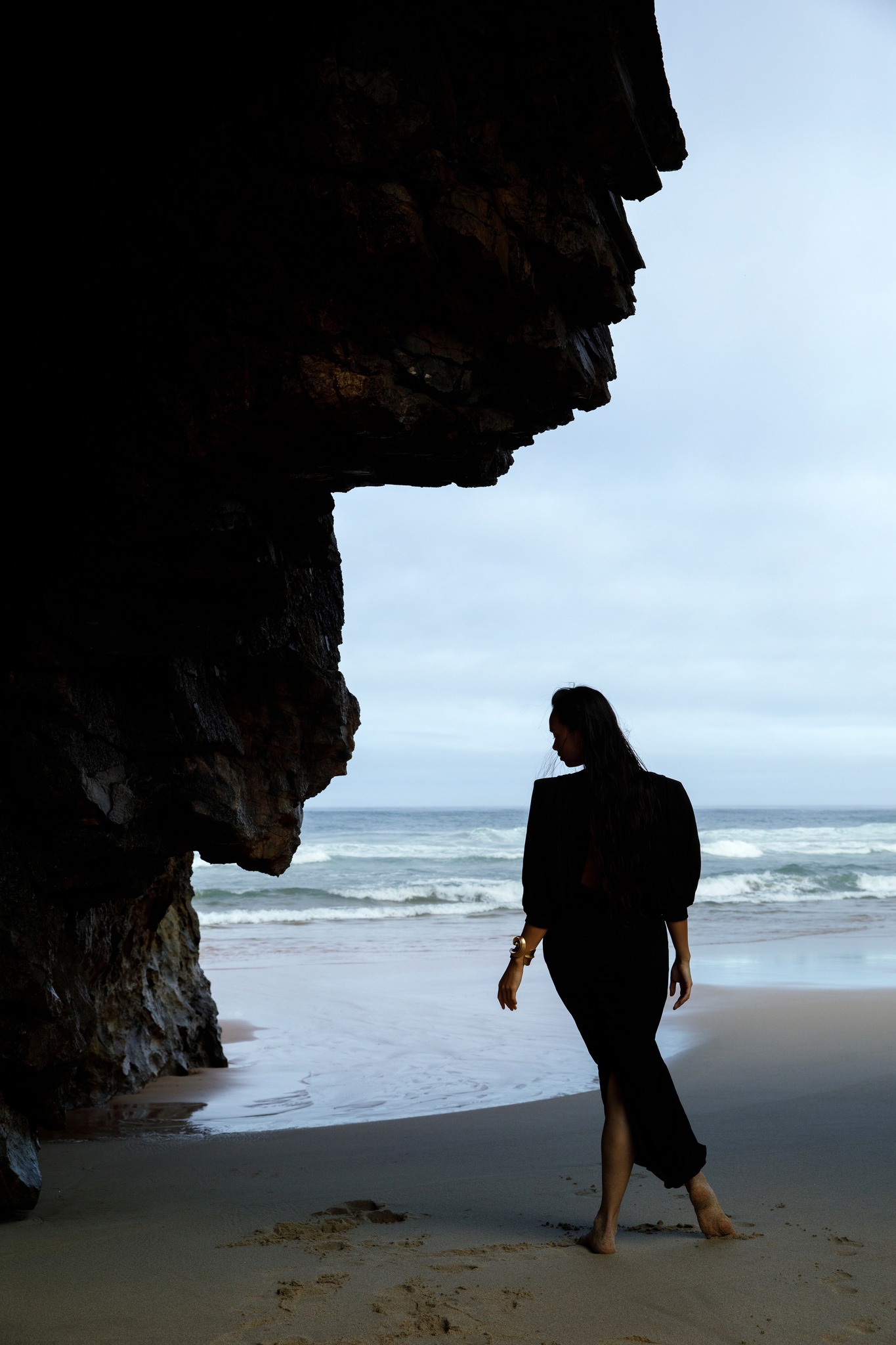 Woman in a black dress standing on a sandy beach next to a dark rocky cliff overlooking the ocean