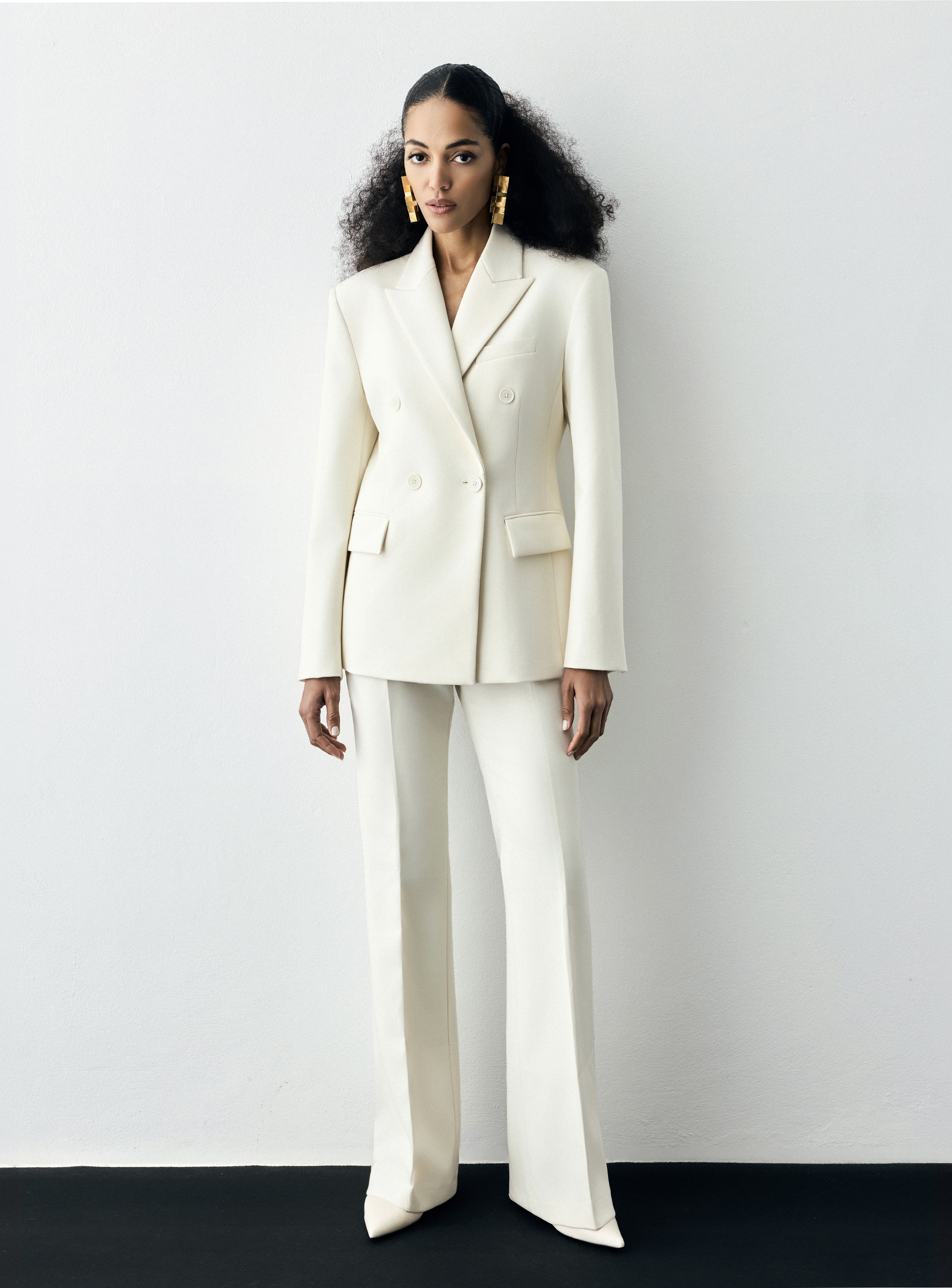 Model wearing a cream double-breasted suit and gold earrings against a white studio background with a dark floor.