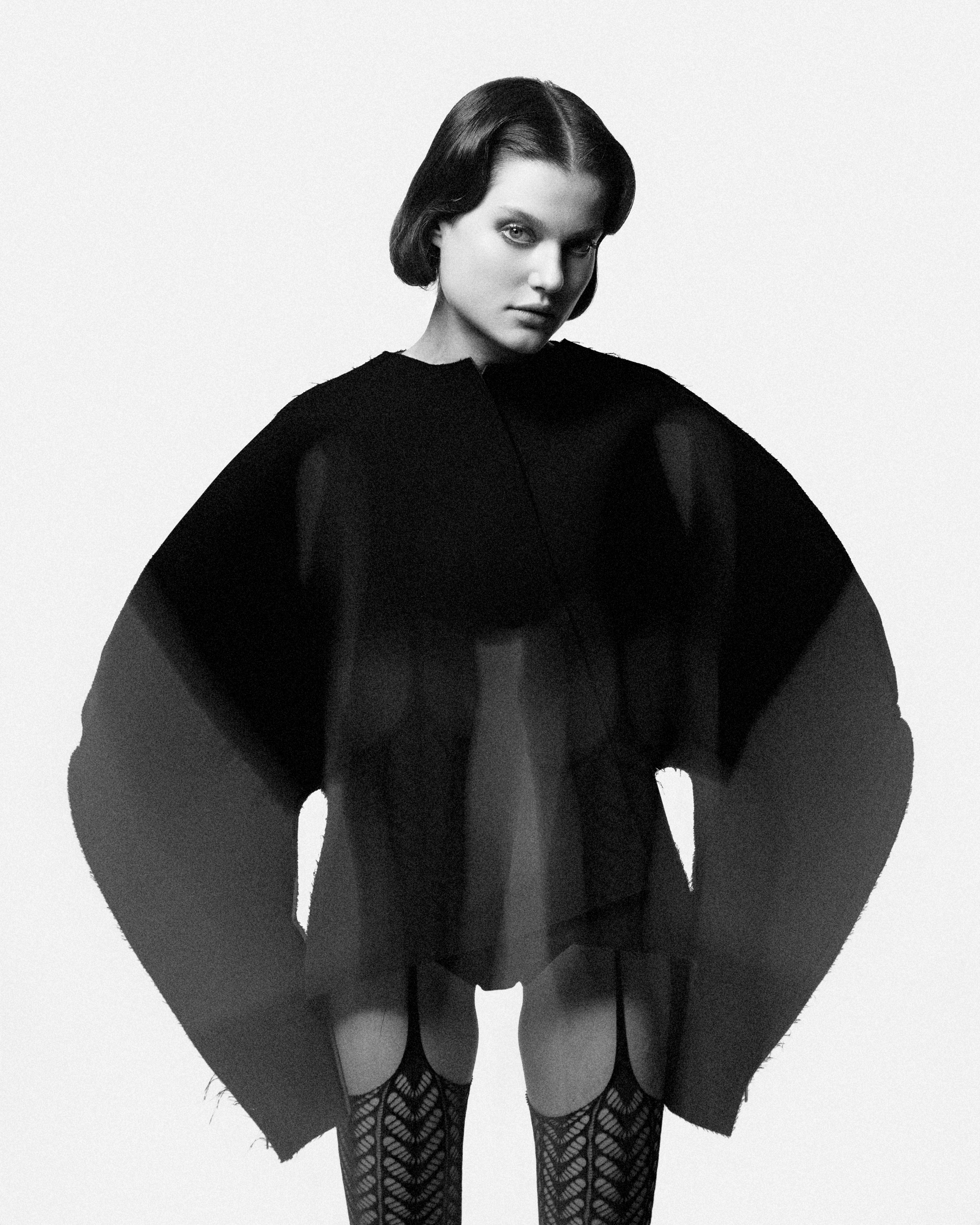 Model posing in a structured black sculptural winged cape jacket and patterned tights against a white studio background.