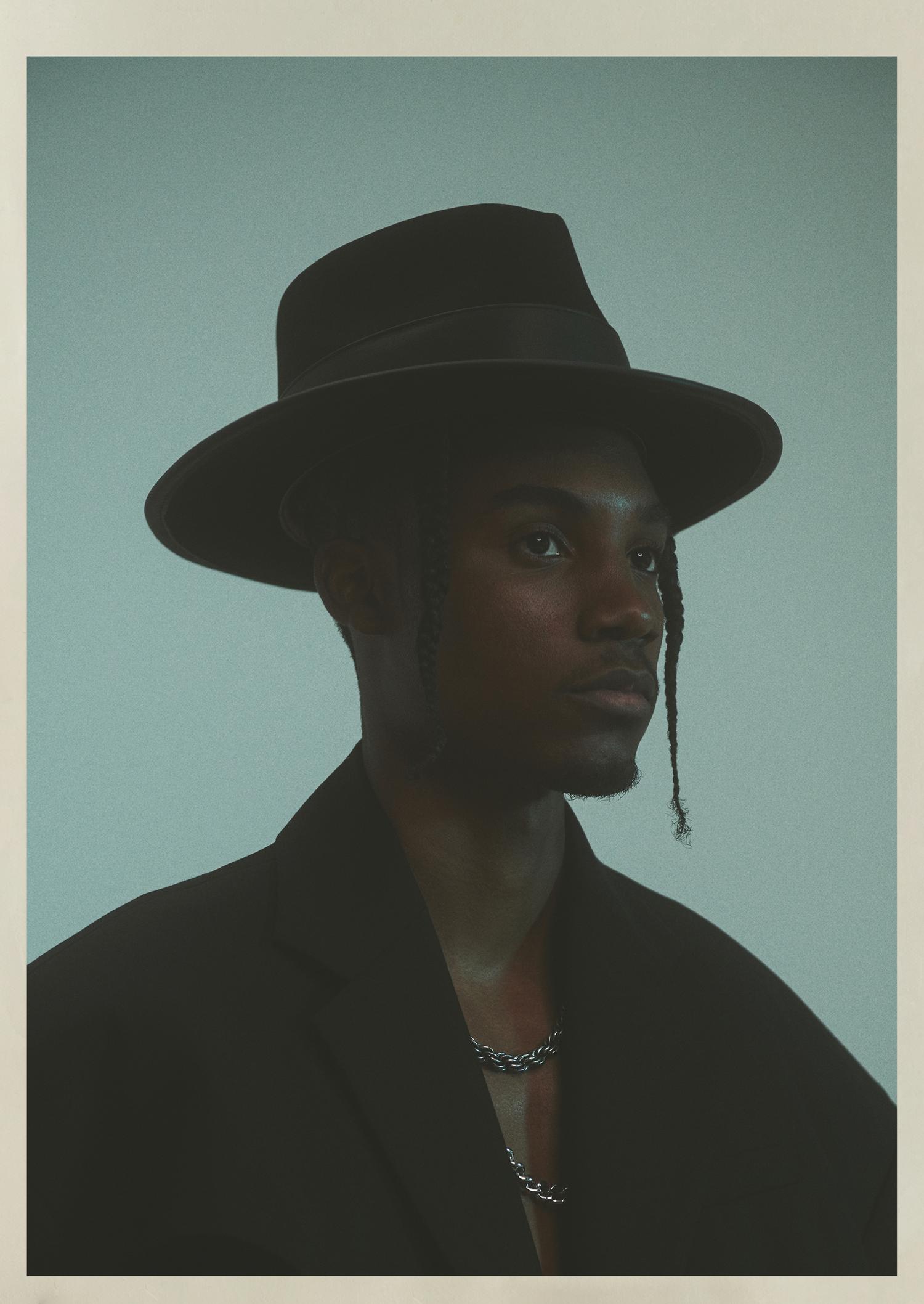 Portrait of a young Black man in a black hat and blazer, wearing a silver chain necklace against a light blue backdrop.