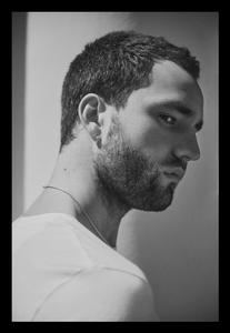 Portrait of a young adult male with short dark hair and a beard, wearing a white t-shirt against a light gray wall in a photo studio.