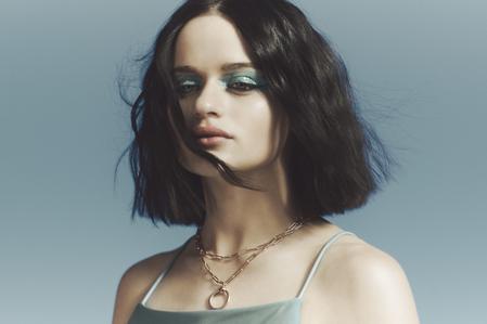 Portrait of a woman with dark hair and blue eye makeup wearing a spaghetti strap top and gold necklaces against a blue background.