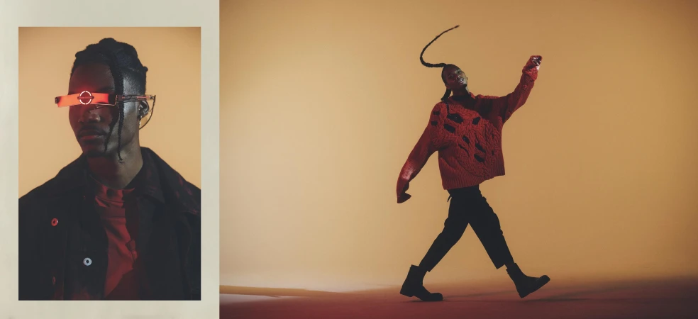 A fashionable Black male model wears a red sweater and cyberpunk glasses in a studio fashion shot with a warm color palette.