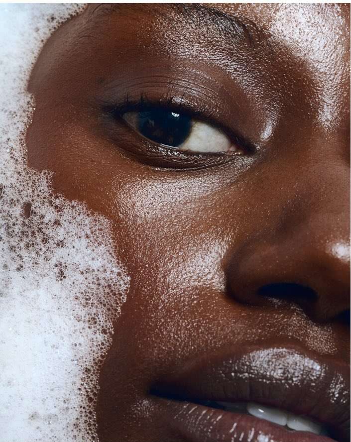 Close-up shot of a woman's face with dark skin, partially covered in white soap bubbles, highlighting her smooth skin and facial features.