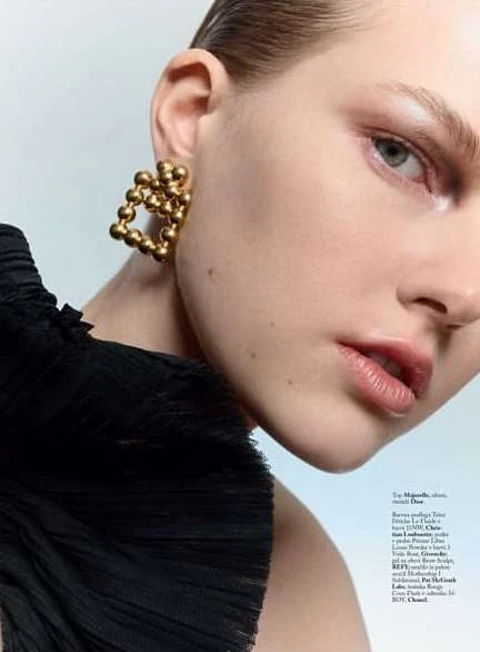 Close-up of a woman with slicked-back hair, wearing a black ruffled top and gold ball-shaped earrings against a light blue background.