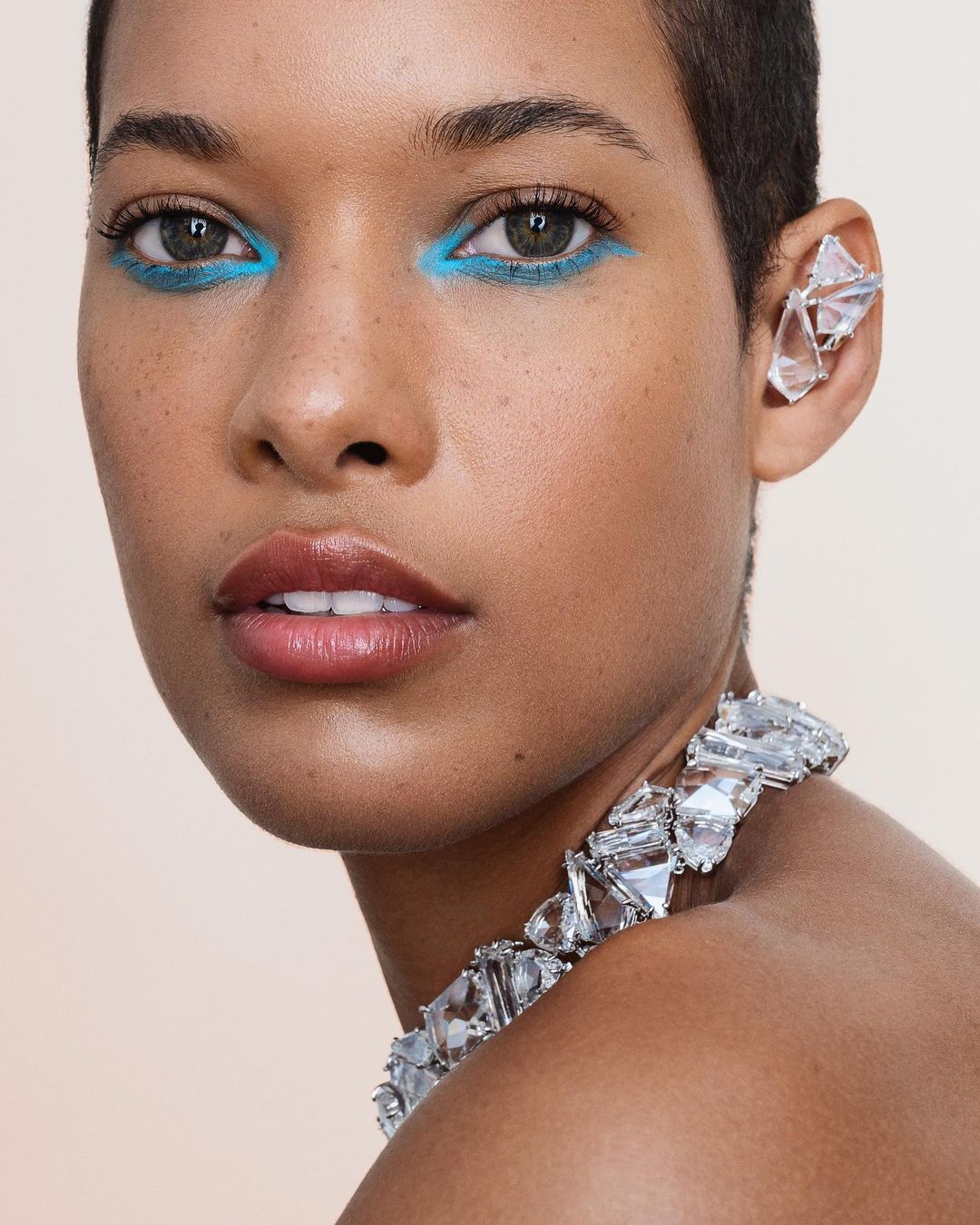 Close-up portrait of a woman with short hair wearing statement crystal jewelry, bright turquoise eyeliner, and a neutral background.