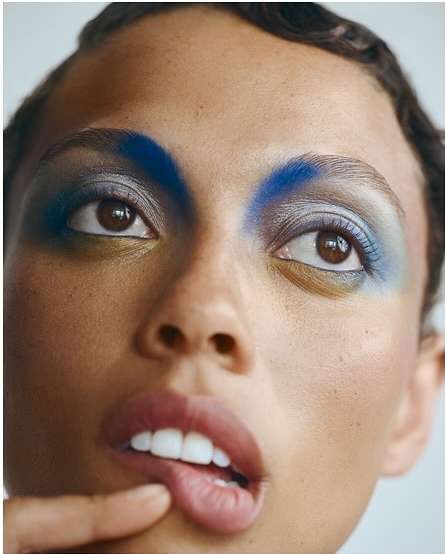 Close-up portrait of a woman with dramatic blue and silver eye makeup, a finger near her lips, and an expression of contemplation.