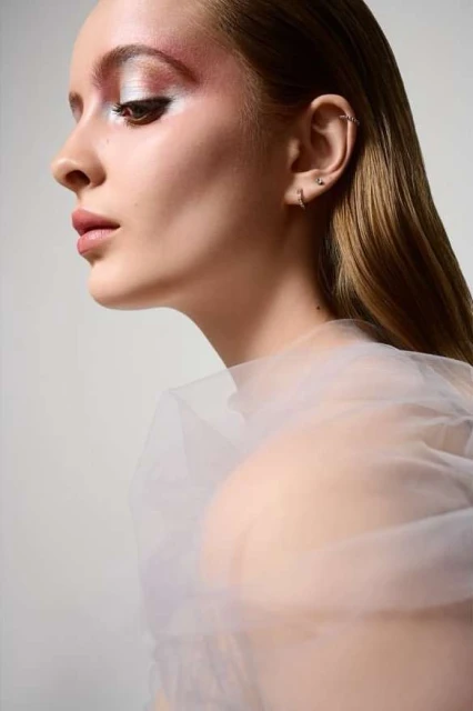 Profile shot of a fair-skinned young woman with silver and pink eye makeup, sheer white fabric and a neutral studio background.