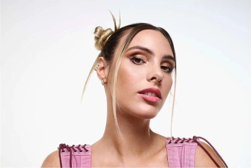Close-up portrait of a young adult woman with blonde-streaked hair in a bun, wearing a pink top against a bright white backdrop.