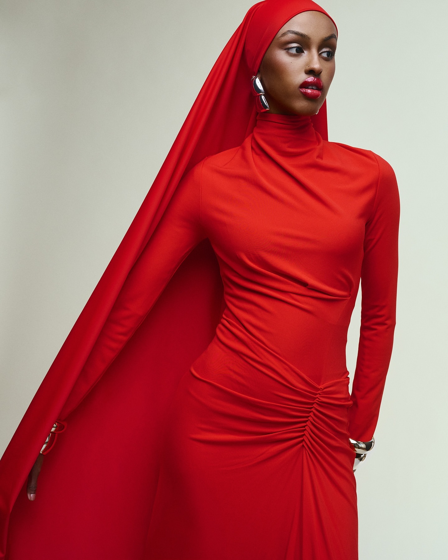 A woman wears a red dress and headscarf, posing in a studio. The garment features long sleeves and draped detailing with silver jewelry.