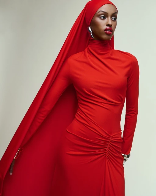 A woman wears a red dress and headscarf, posing in a studio. The garment features long sleeves and draped detailing with silver jewelry.