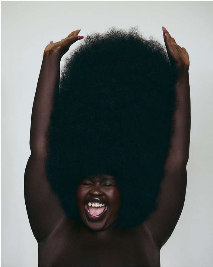 Portrait of an adult female with a very large afro, mouth open, hands raised, against a pale grey background in a studio setting.