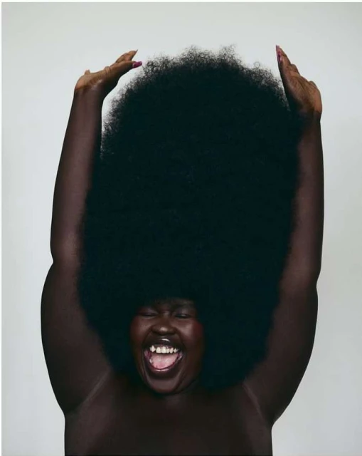 Portrait of an adult female with a very large afro, mouth open, hands raised, against a pale grey background in a studio setting.