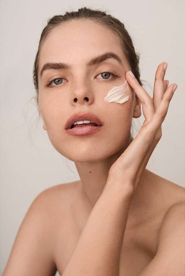 Portrait of a young woman applying cream to her face, beauty shot, skincare routine, close-up on neutral background.
