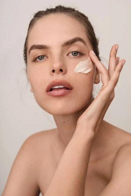Portrait of a young woman applying cream to her face, beauty shot, skincare routine, close-up on neutral background.