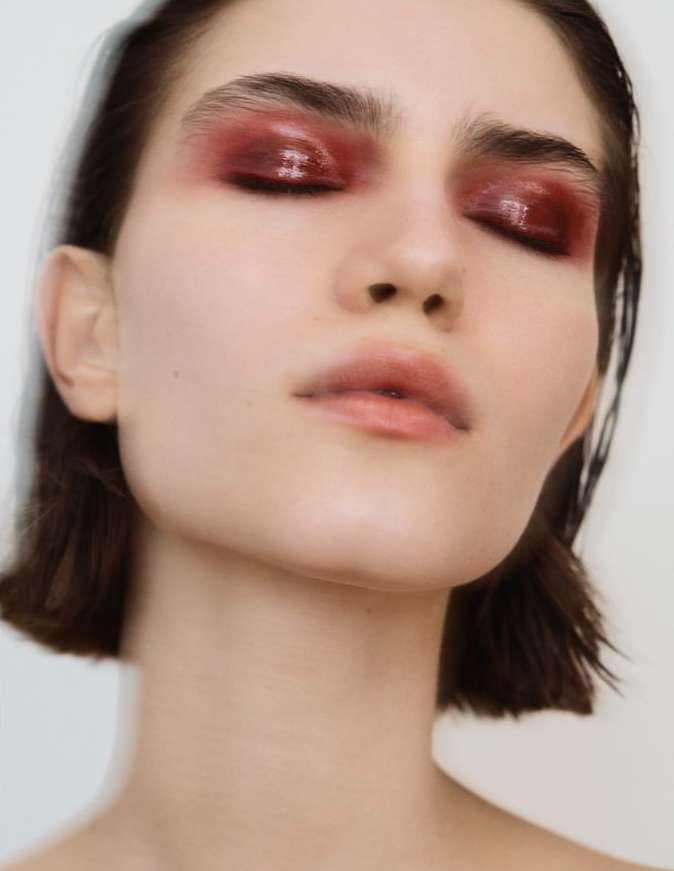 Close-up portrait of a young fair-skinned woman with bold glossy red eye makeup and dark hair, set against a blurred light background.