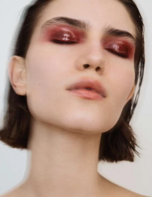 Close-up portrait of a young fair-skinned woman with bold glossy red eye makeup and dark hair, set against a blurred light background.