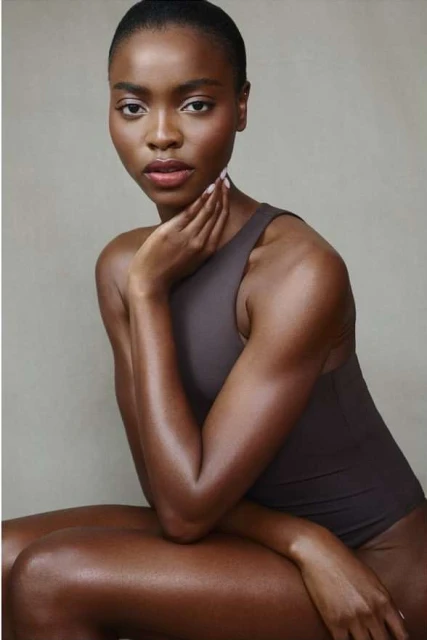 Portrait of a young African American woman in a studio. She is seated, wearing a brown sleeveless leotard, her hand gently touching her neck.