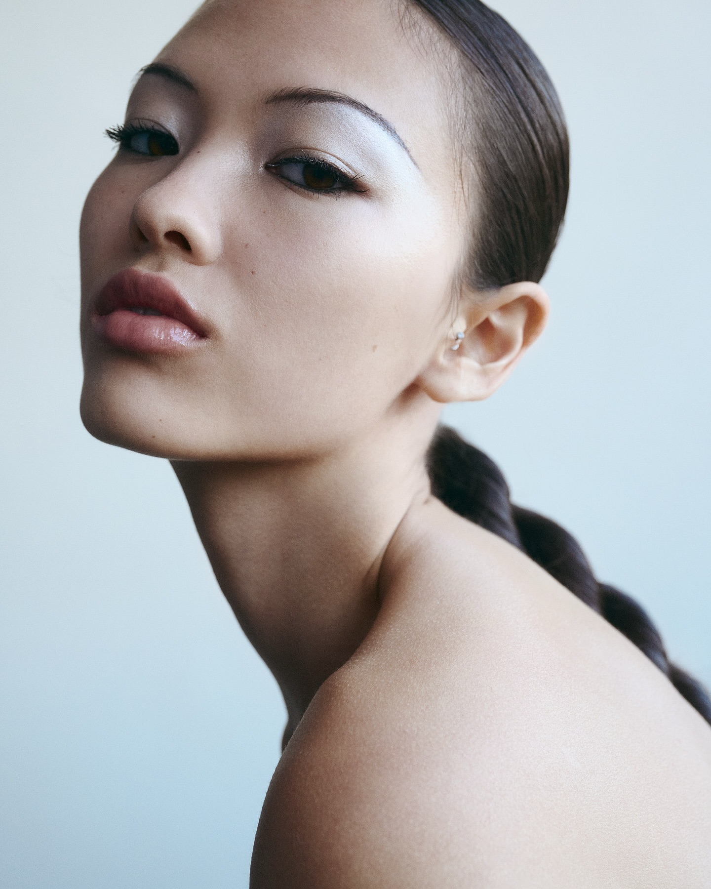 Close-up studio shot of a young adult Asian woman with braided hair, silver eyeshadow, and light pink lipstick against a light blue background.