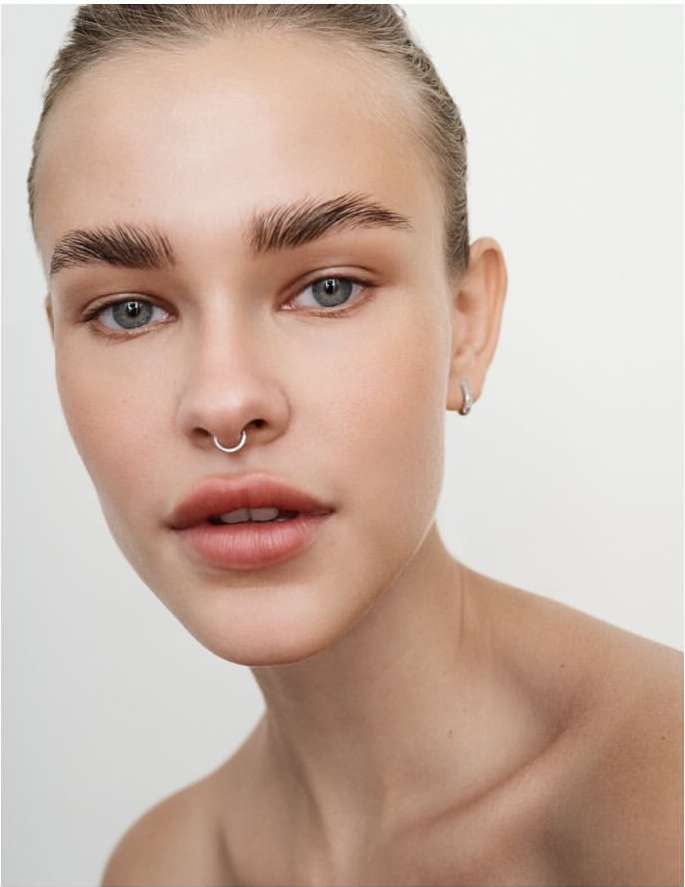Close-up shot of a young, light-skinned woman with a nose ring and light makeup posing in front of a plain light-colored background.