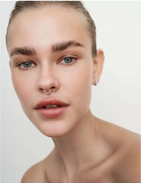 Close-up shot of a young, light-skinned woman with a nose ring and light makeup posing in front of a plain light-colored background.