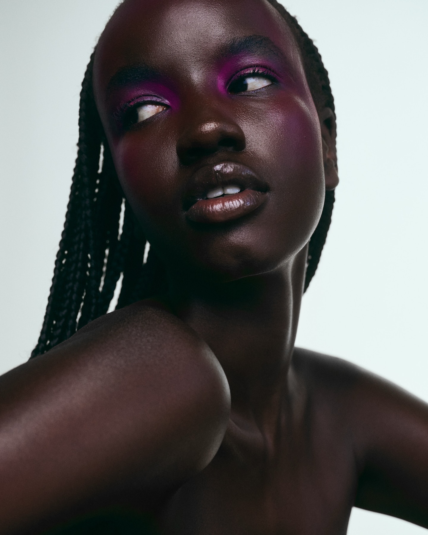 Close-up portrait of a woman with braided hair and artistic purple makeup against a bright white background in a beauty studio shoot.