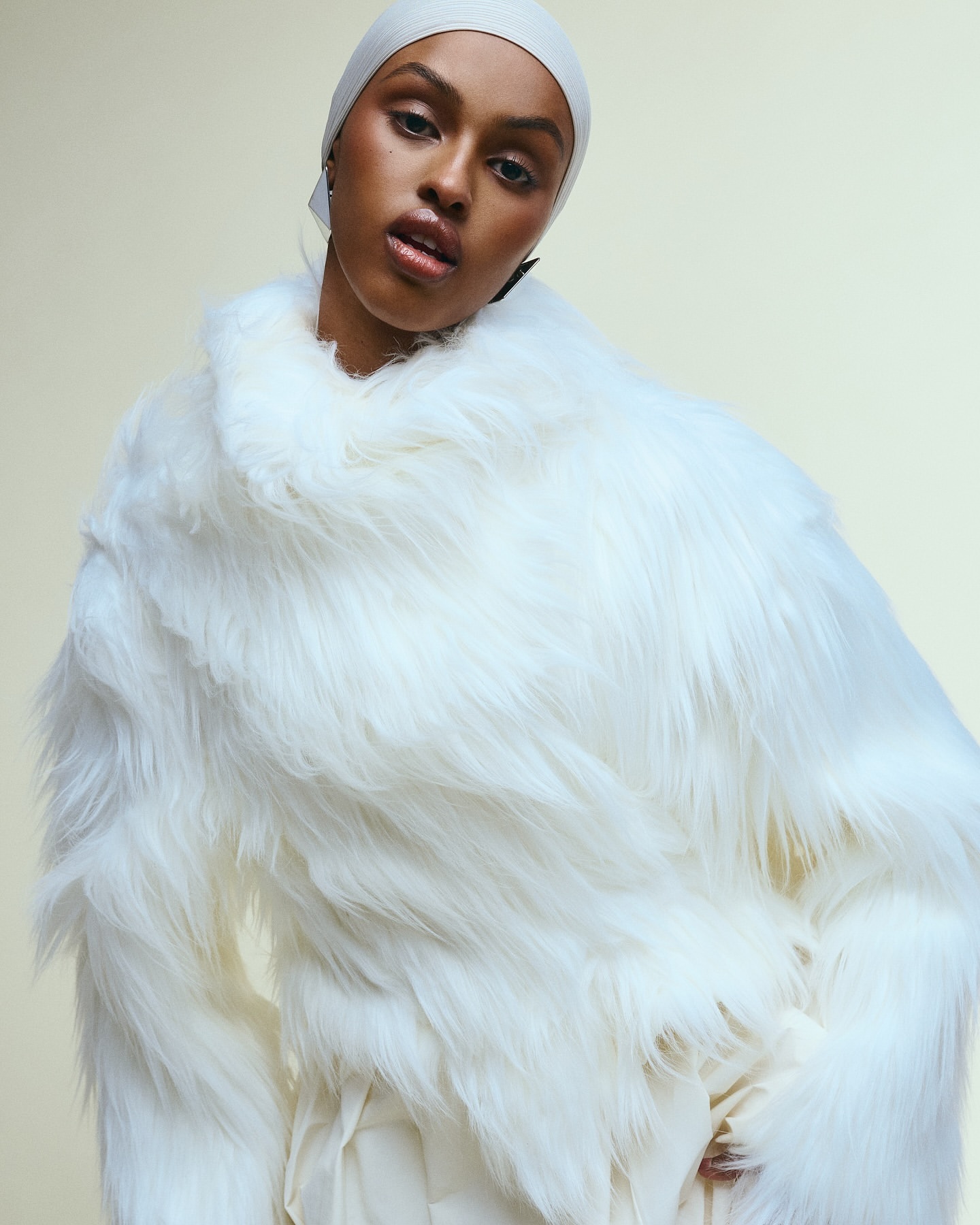 Fashion portrait of a Black female adult wearing a white headscarf and a fur coat with a neutral background in an indoor setting.