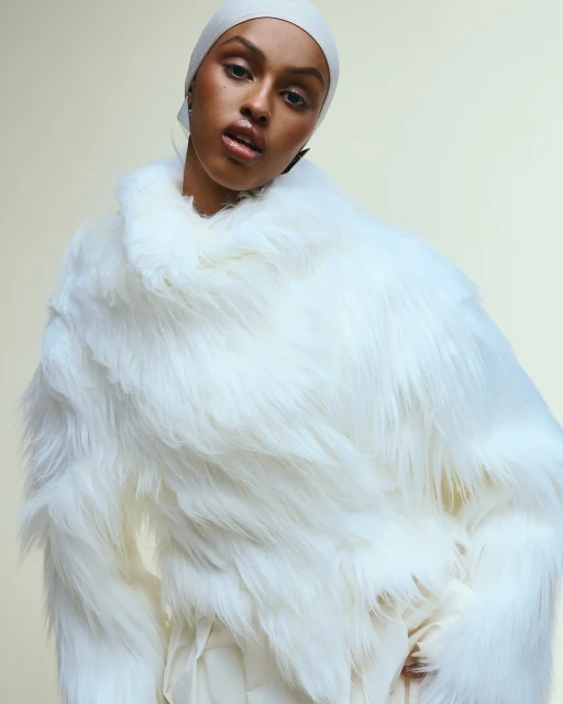 Fashion portrait of a Black female adult wearing a white headscarf and a fur coat with a neutral background in an indoor setting.