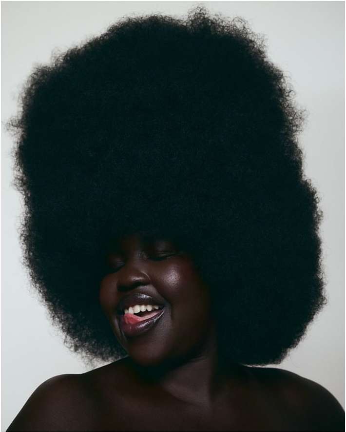 Close-up portrait of an African American woman with large afro hairstyle, glossy lips, and a joyful expression in front of a plain white background.