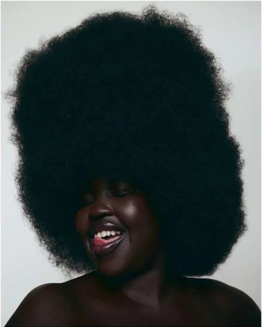 Close-up portrait of an African American woman with large afro hairstyle, glossy lips, and a joyful expression in front of a plain white background.