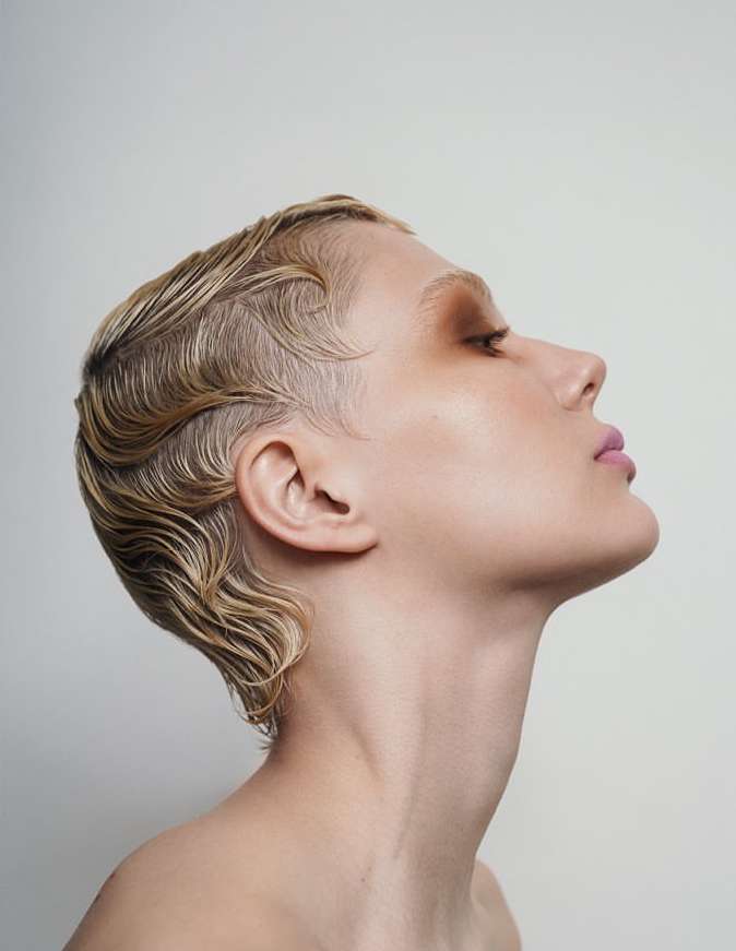Close-up profile of a woman with short, blonde, wavy hair, light skin, and light pink lipstick against a plain white background.