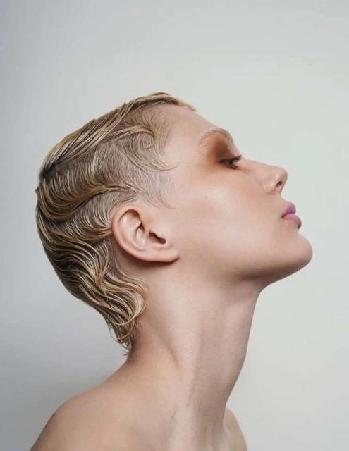 Close-up profile of a woman with short, blonde, wavy hair, light skin, and light pink lipstick against a plain white background.