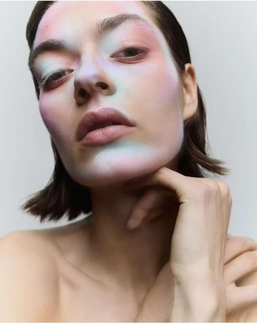 Close-up portrait of a young adult woman with light skin, colorful makeup, and short brown hair, holding her chin, in a beauty studio.